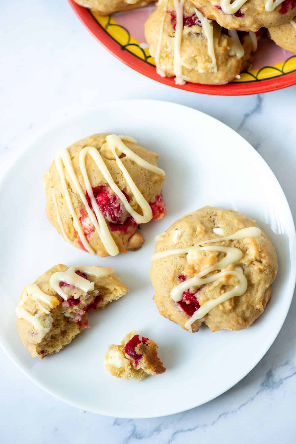 Overhead photo of two white chocolate cranberry orange cookies and some cookie crumbs on a white plate in front of a pink plate with more cookies