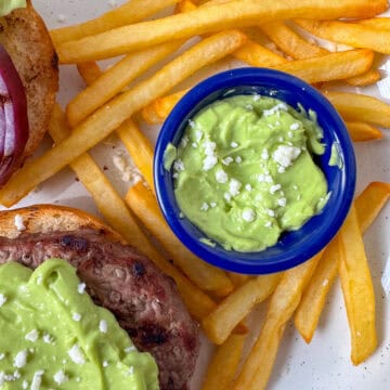 Overhead photo of a sauce cup filled with avocado aioli surrounded by French fries and a view of a partial burger topped with avocado aioli