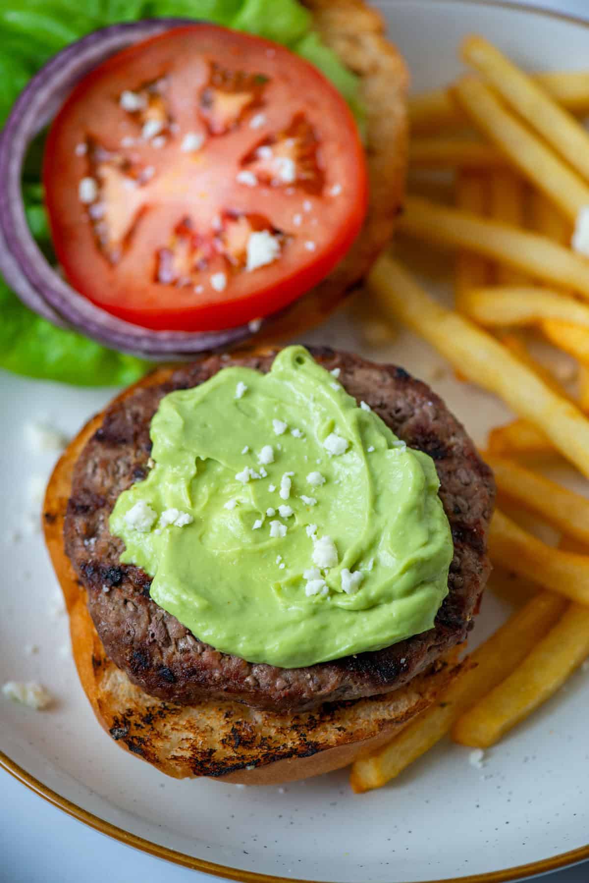 Overhead photo of a burger with avocado aioli sauce on top with tomatoes, onions, lettuce and French fries in the background