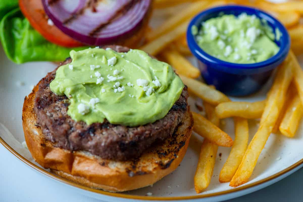 Overhead angled view of open face burger topped with avocado aioli surrounded by fries and a condiment cup filled with avocado aioli