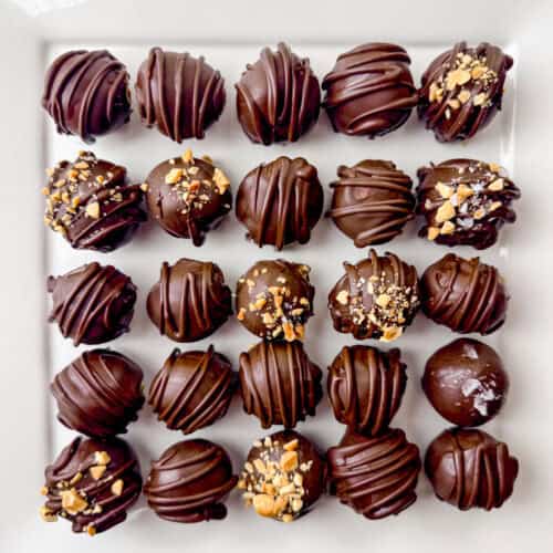 Overhead photo of 25 chocolate peanut butter truffles on a square, white plate. Some are decorated with crushed peanuts.