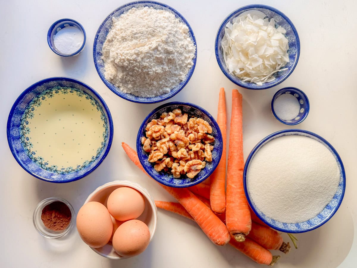 Ingredients needed to make carrot bundt cake: all purpose flour, white sugar, neutral vegetable oil, eggs, carrots, walnuts, baking soda, salt, and coconut to toast.