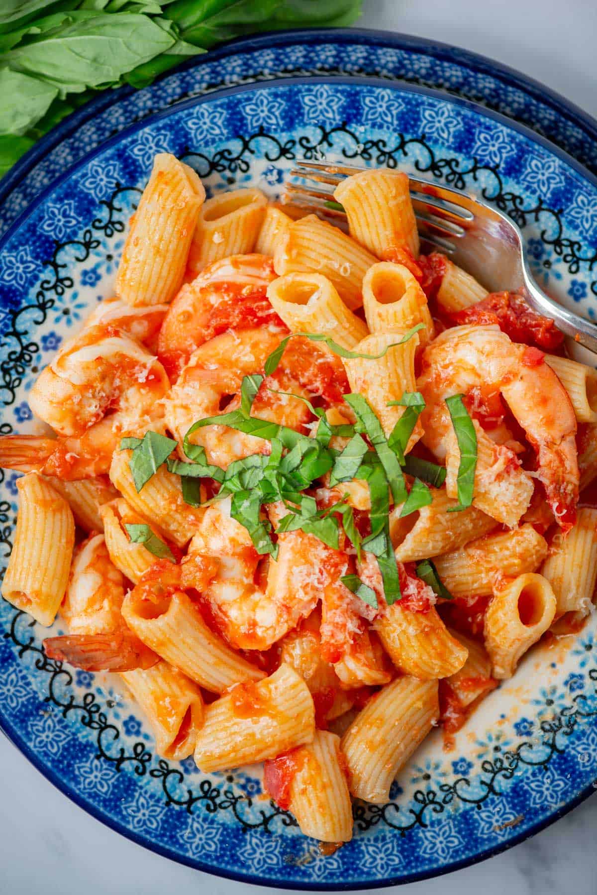 Overhead view of shrimp rigatoni with tomato butter sauce topped with parmesan cheese and a basil garnish with more basil in the background.