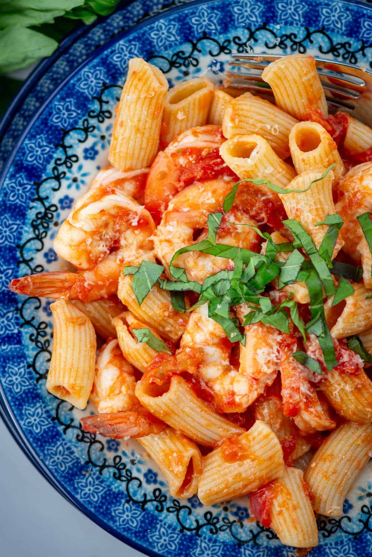Overhead view of a partial plate of rigatoni with shrimp and tomato butter sauce with a basil garnish on a blue Polish pottery plate. 