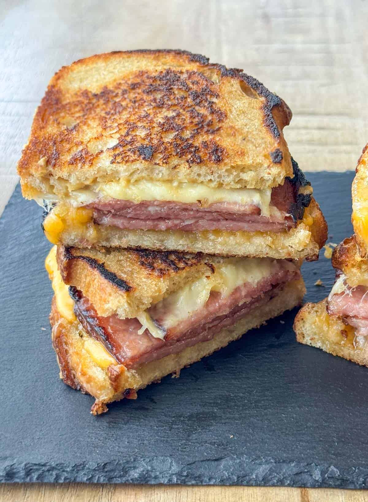 Side view photo of a grilled ham and cheese sandwich that's been cut in half and stacked on top of each other with a partial sandwich visible to the right