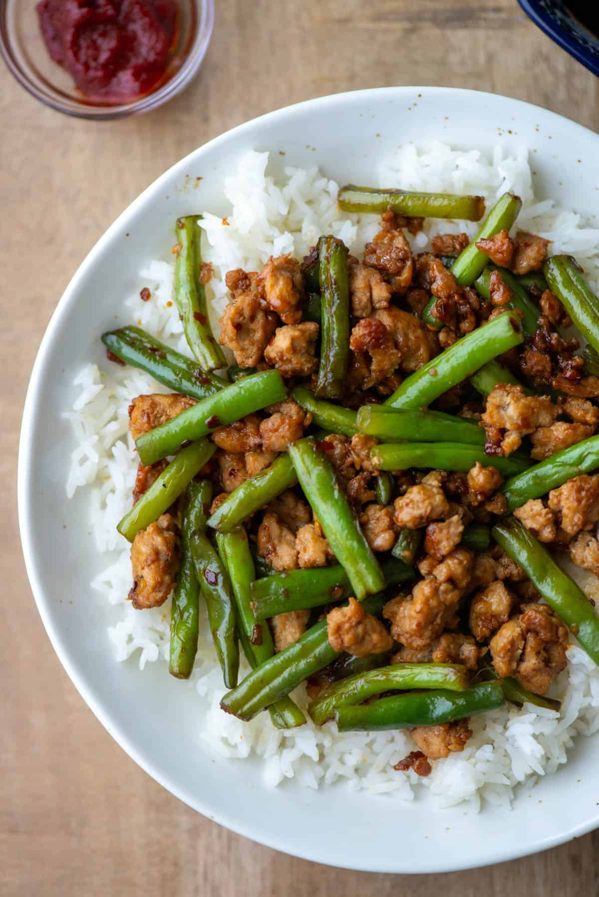 Overhead view of a partial plate of ground chicken stir fry with green over white jasmine rice with a small bowl of Gochujang sauce in the background