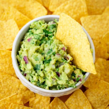 A bowl of guacamole with a chip sticking out surrounded by chips on a plate