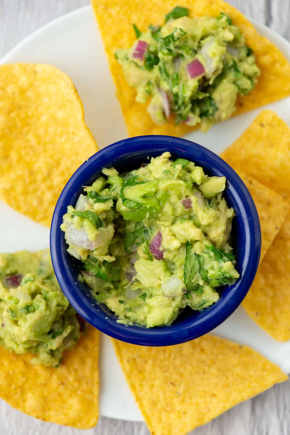 Guacamole in a small blue bowl surrounded by tortilla chips on a plate, some with guacamole on them