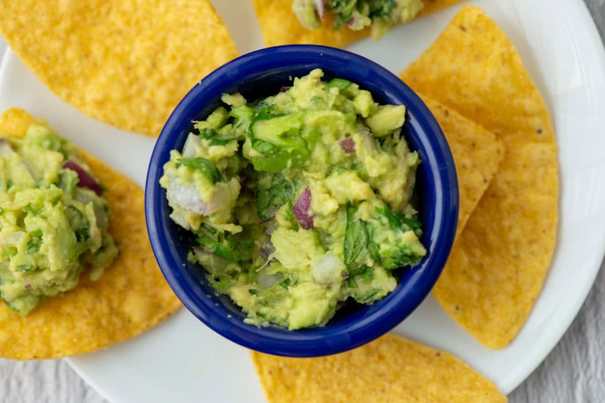 Guacamole in a small blue bowl surrounded by tortilla chips on a plate, some with guacamole on them