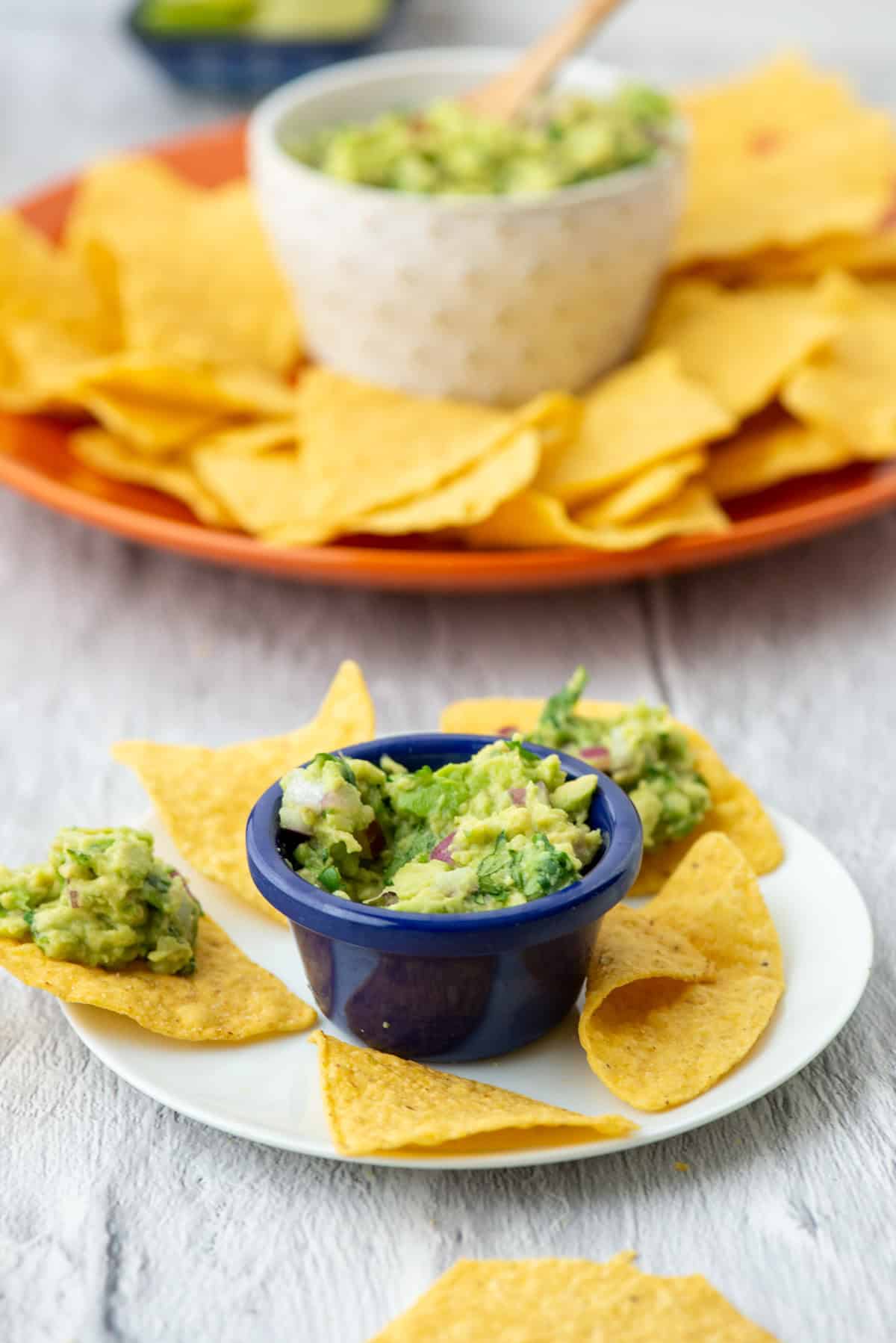 A small bowl of guacamole on a small plate with tortilla chips in front of a larger bowl of guacamole on an organ plate surrounded by more tortilla chips
