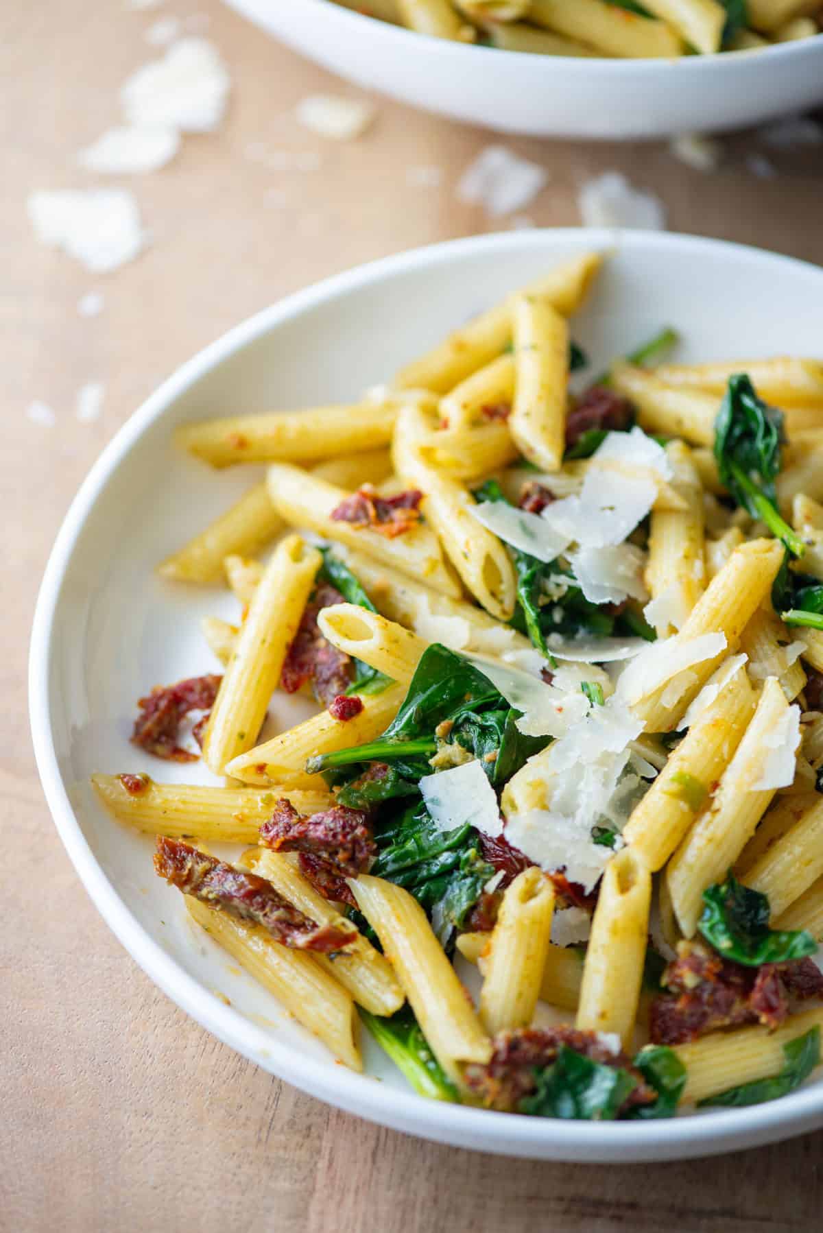 Angled overhead photo of pasta pesto with sun-dried tomatoes on a plate with a serving bowl filled with the same pasta in the background
