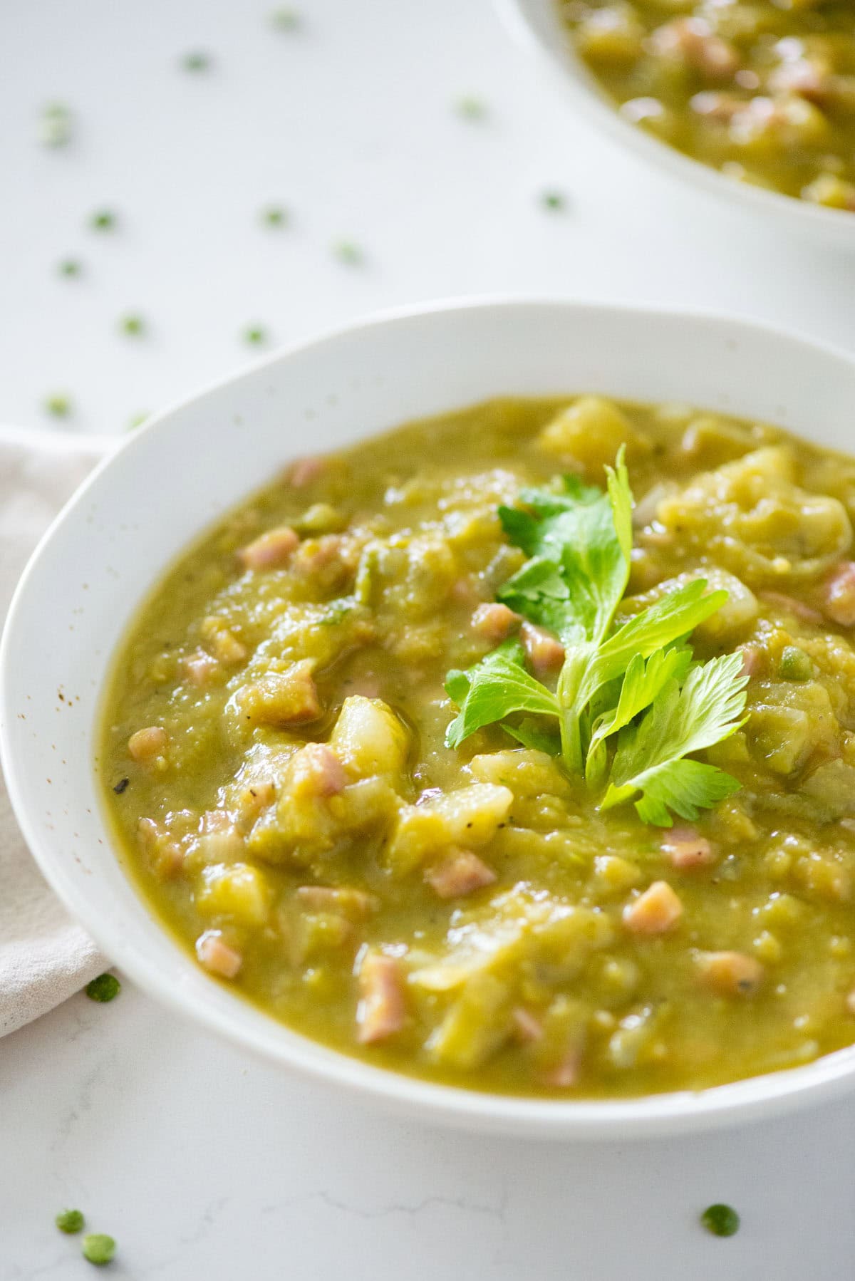 Angled view of split pea soup garnished with celery leaves with another bowl and uncooked split peas scattered on the table