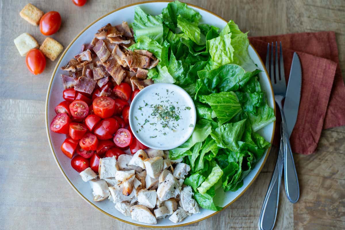 Overhead view of a Chicken BLT Salad with ranch dressing in the middle and a fork, knife and napkin on the side with loose grape tomatoes and croutons in the background
