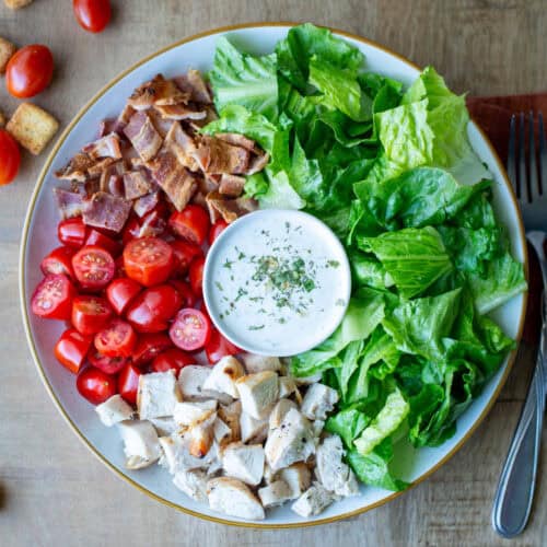 Overhead view of a Chicken BLT Salad with ranch dressing in the middle and a fork, knife and napkin on the side with loose grape tomatoes and croutons in the background