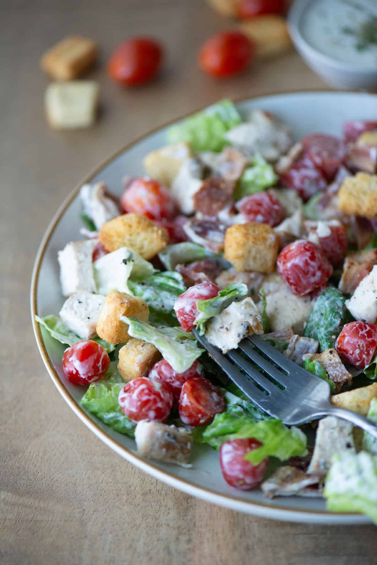 Angled view of Chicken BLT Salad with ranch dressing mixed in and topped with croutons with a fork in the middle and loose tomatoes and croutons in the background