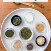 Overhead view of dried spices for ranch seasoning mix in little bowls on a white plate: parsley, minced onion, dill, onion powder, chives, garlic powder, salt and pepper with two measuring spoons to the side with a title overlay that says Dairy Free Ranch Seasoning Mix