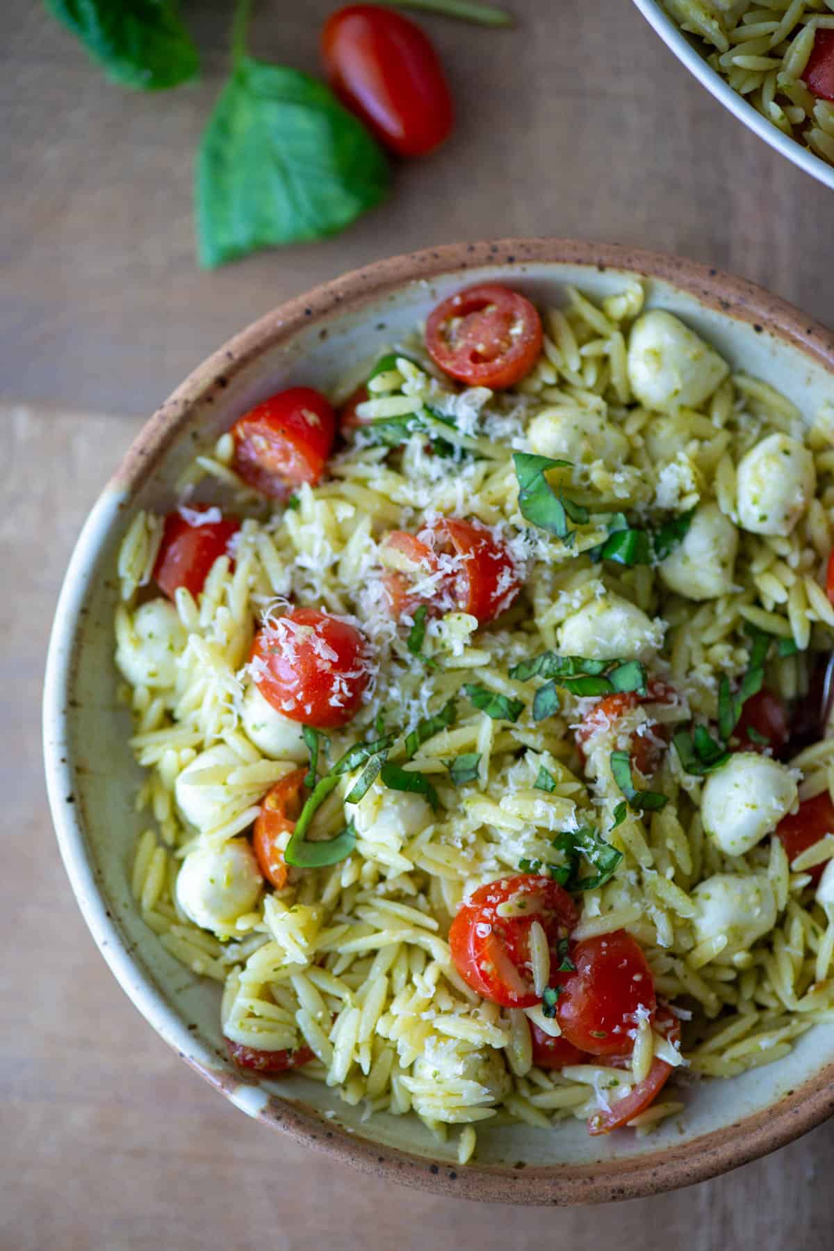 Overhead view of pesto orzo pasta salad on a plate garnished with basil and parmesan cheese with some loose tomatoes and basil in the background 