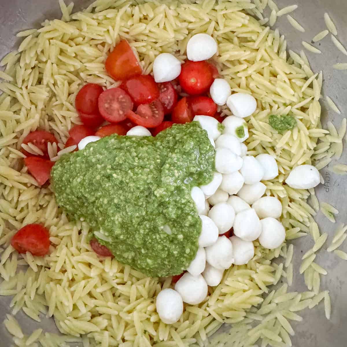 Orzo pasta with mozzarella pearls, sliced cherry tomatoes and basil pesto in a bowl before it's been mixed.