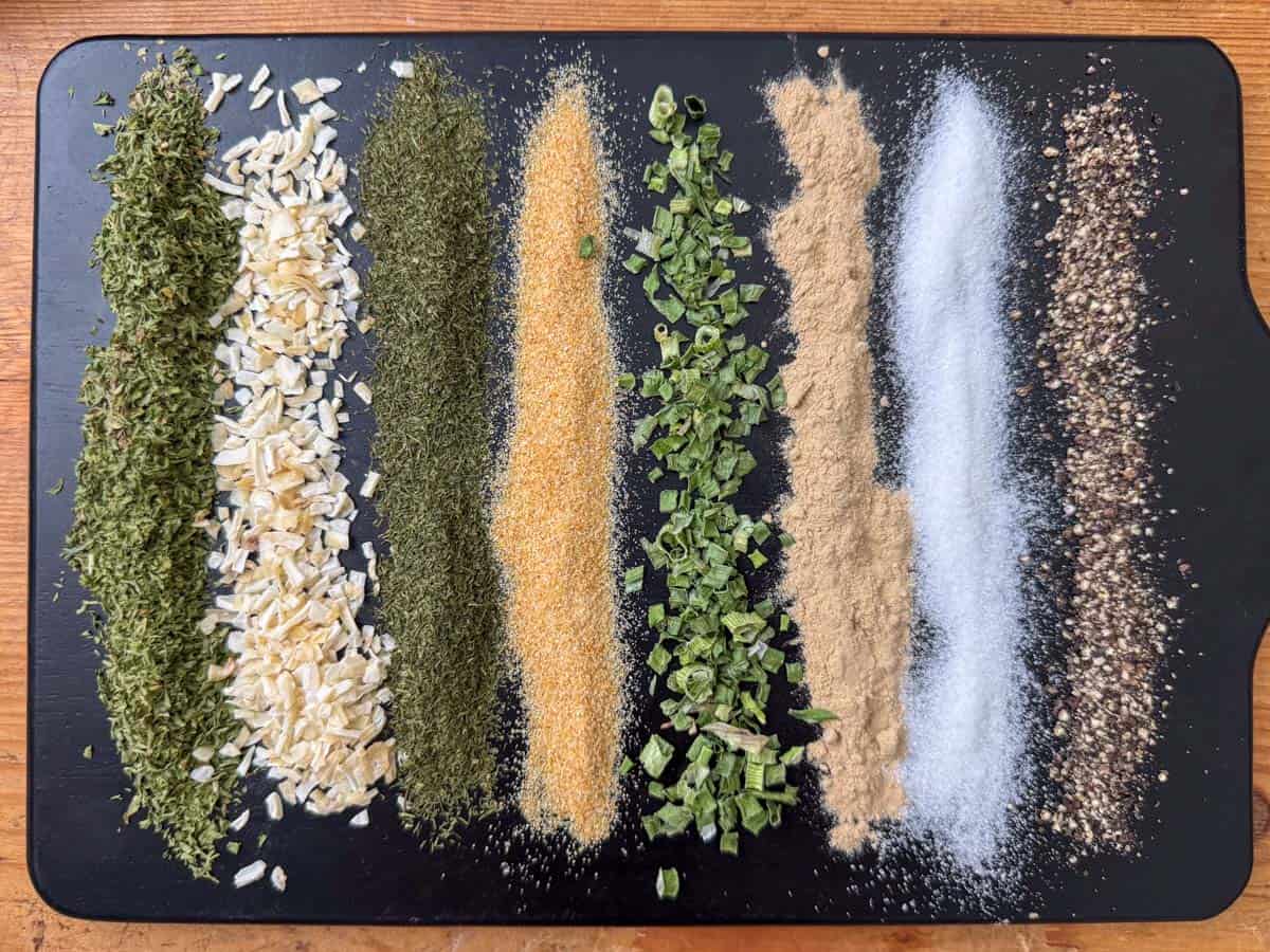 Overhead view of dried spices for ranch seasoning mix in lines on a cutting board: parsley, minced onion, dill, onion powder, chives, garlic powder, salt and pepper