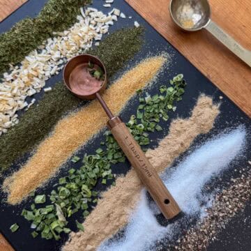 Overhead view of dried spices for ranch seasoning mix in lines on a cutting board: parsley, minced onion, dill, onion powder, chives, garlic powder, salt and pepper with a measuring spoon on top and one to the side
