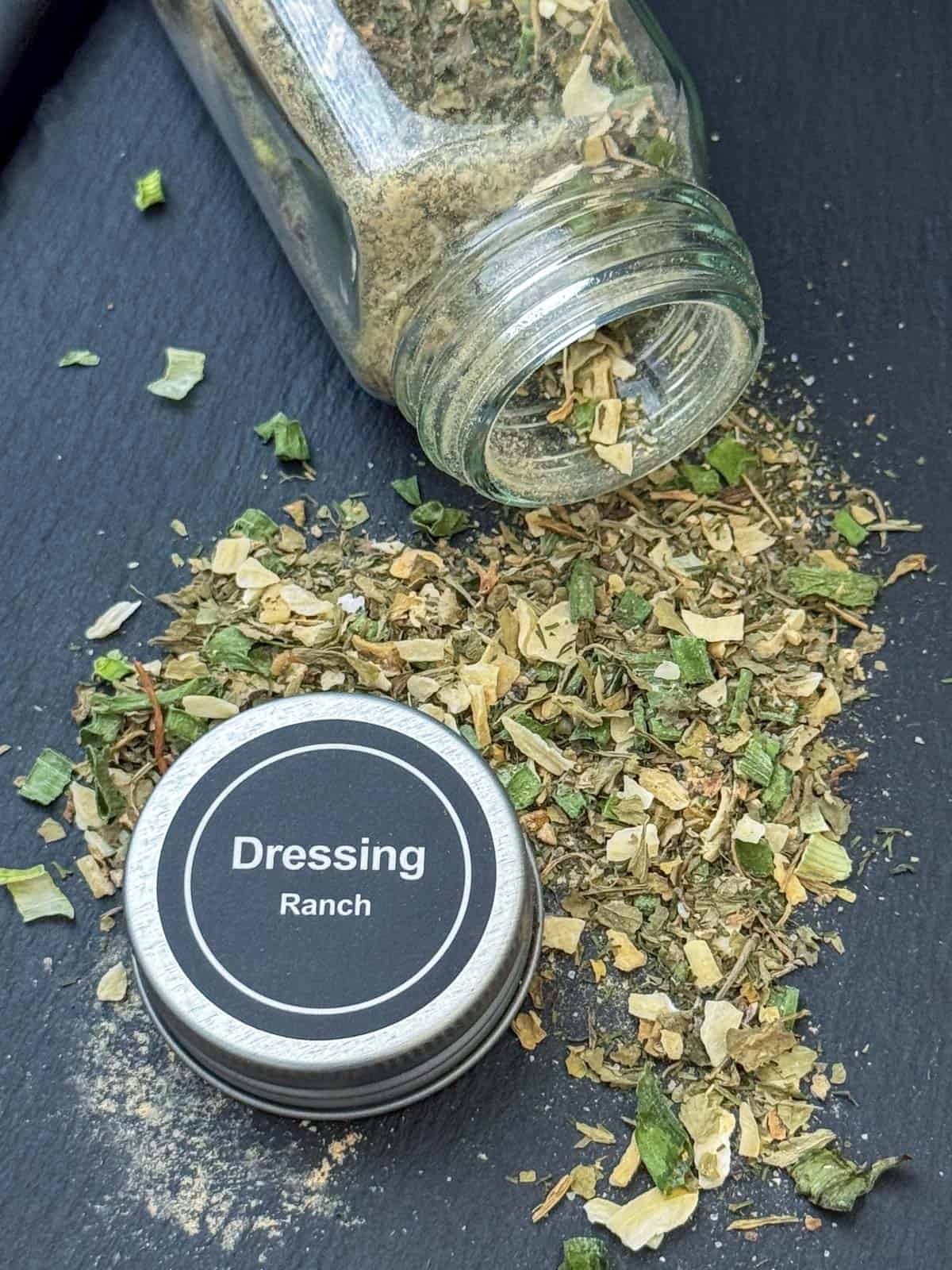 Overhead view of a jar of dried ranch seasoning mix spilling out on to a cutting board with the jar cap that says Dressing Ranch on top in the middle of the dried spices
