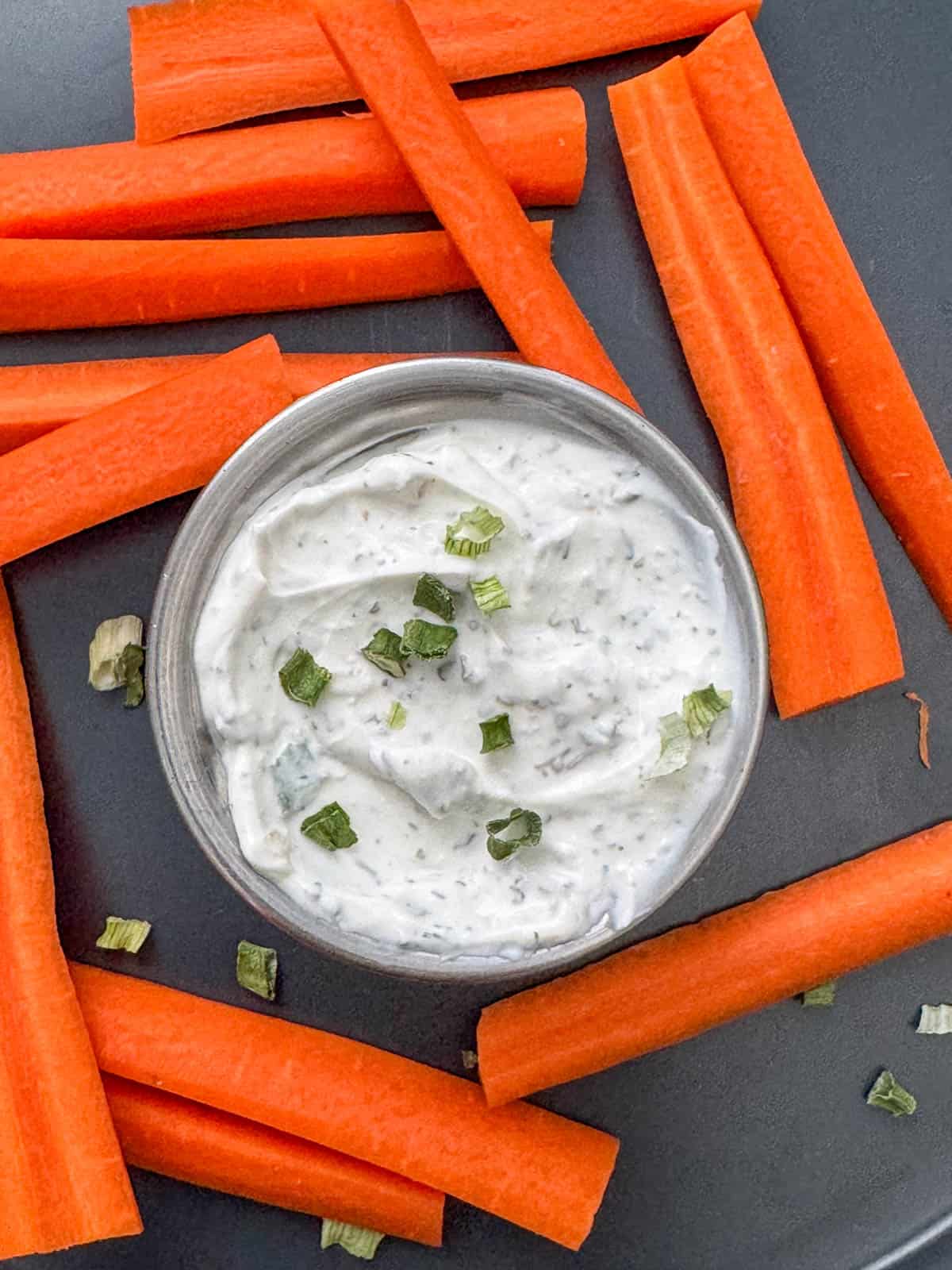 Overhead view of a little bowl of ranch on a plate with cut carrots and dried chives sprinkled around