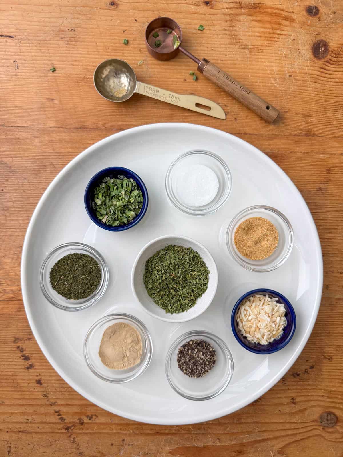 Overhead view of dried spices for ranch seasoning mix in little bowls on a white plate: parsley, minced onion, dill, onion powder, chives, garlic powder, salt and pepper with two measuring spoons to the side