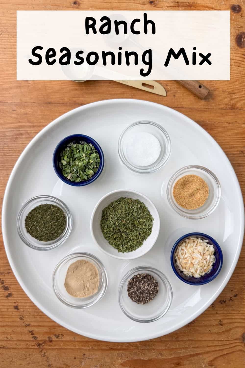 Overhead view of dried spices for ranch seasoning mix in little bowls on a white plate: parsley, minced onion, dill, onion powder, chives, garlic powder, salt and pepper with a two measuring spoons to the side with a title overlay that says Ranch Seasoning Mix