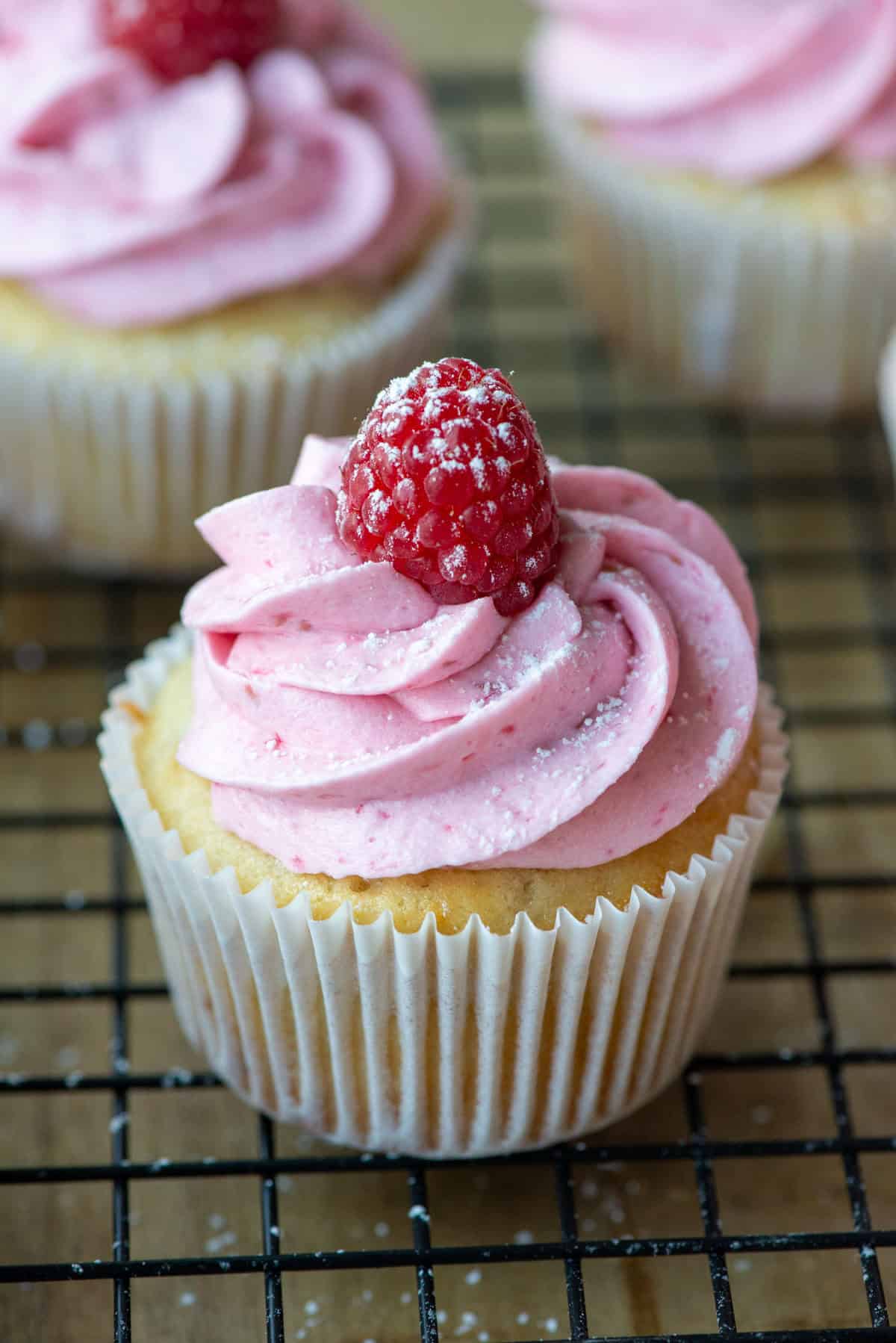A single raspberry white chocolate cupcake on a cooling rack with two other cupcakes in the background