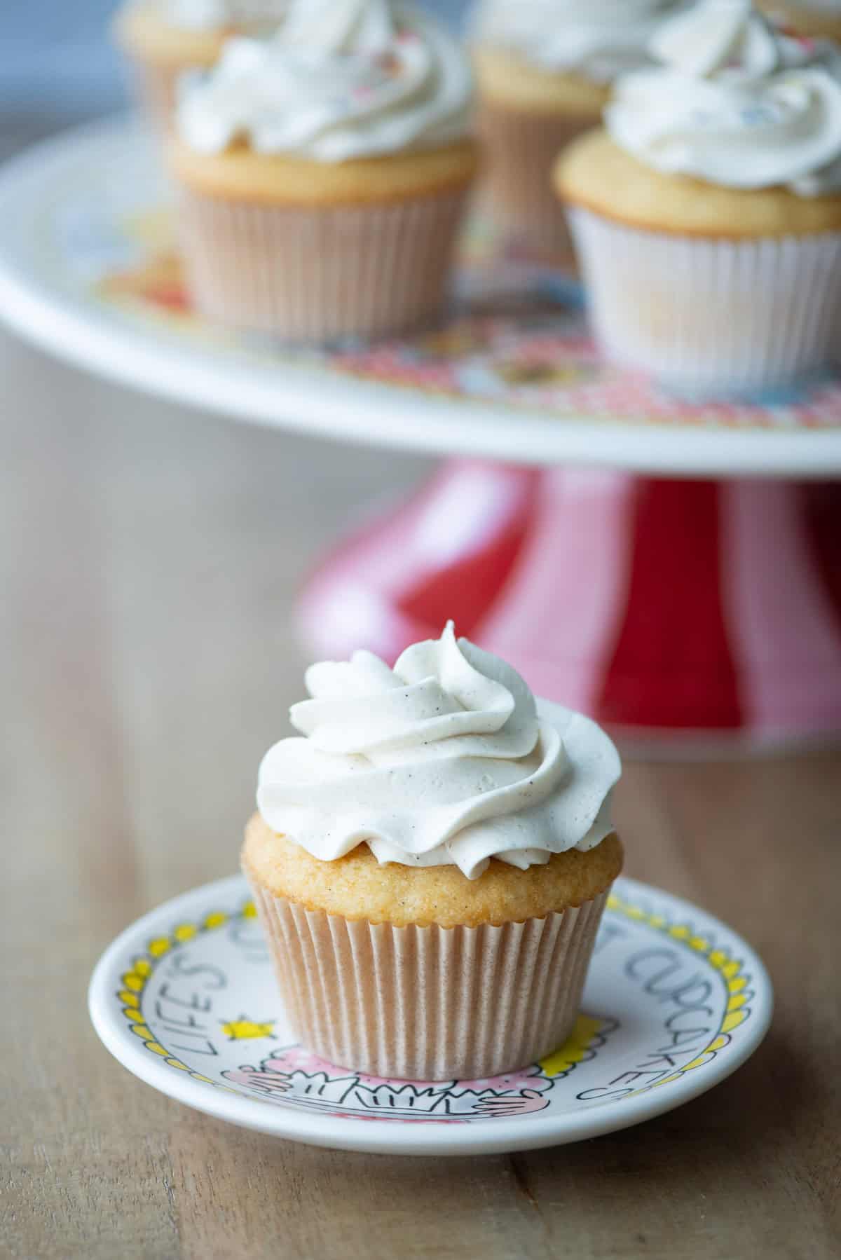 Side view of one cupcake on a small plate in front of a cake stand filled with more cupcakes.