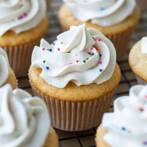 Close up view focused on one vanilla bean cupcake with vanilla bean buttercream with sprinkles surrounded by other cupcakes in partial view