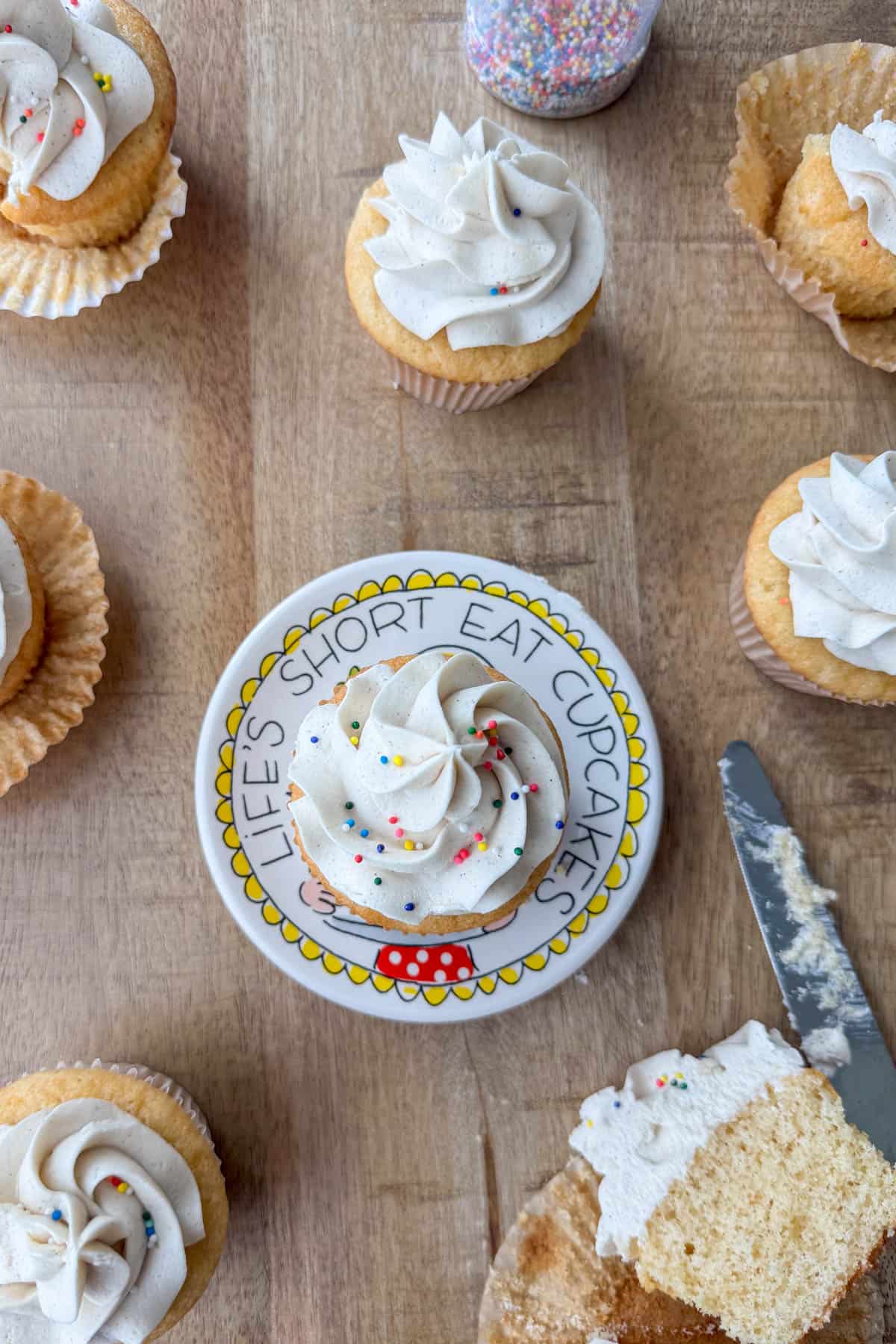 Overhead view of a vanilla bean cupcake on a small plate that says 'LIFE'S SHORT EAT CUPCAKES' surrounded by other cupcakes on the table in different states, some with the wrapper pealed, one with a bite taken out and one cut in half with a knife next to it