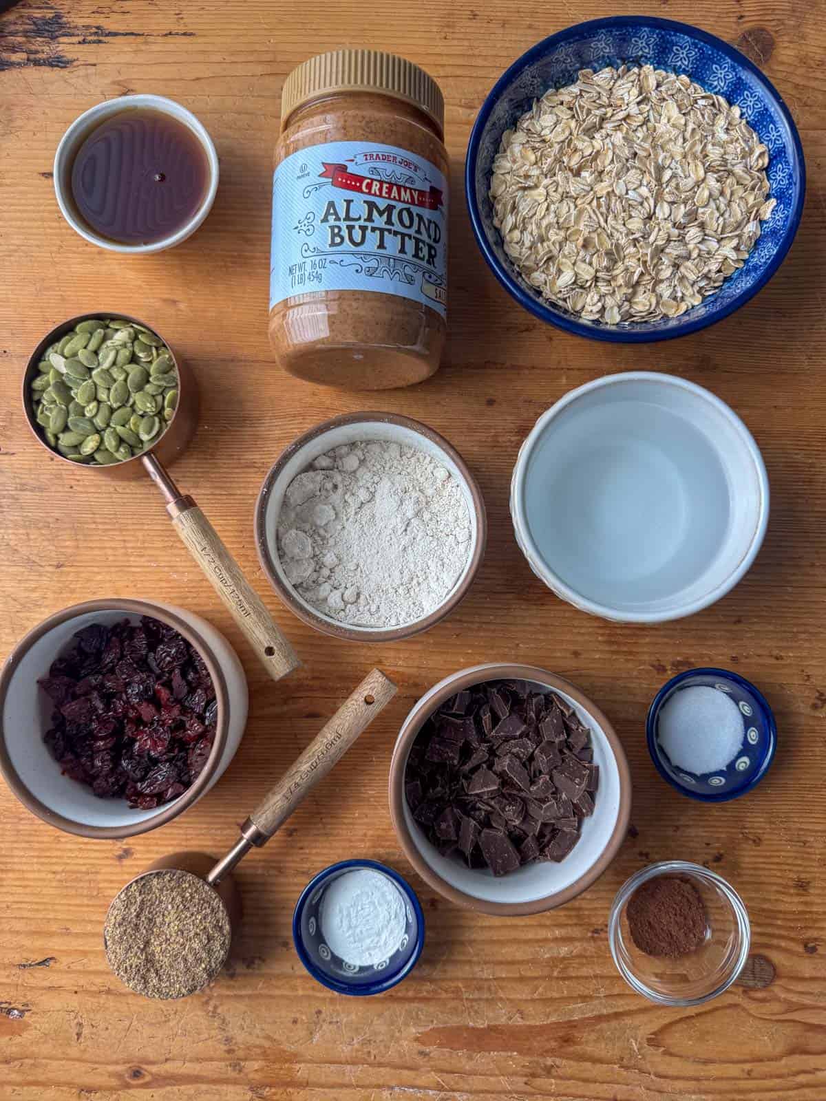 Overhead view of ingredients needed to make oatmeal breakfast cookies: oatmeal, almond butter, maple syrup, pumpkin seeds, oat flour, water, flaxseed meal, dried cherries, chocolate chunks, baking powder, cinnamon and salt.
