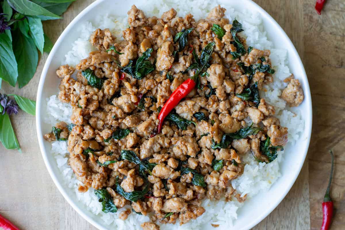 Overhead view of a full plate of Thai Basil Chicken over a bed of white rice with Thai Basil and Thai red chilies visible on the table