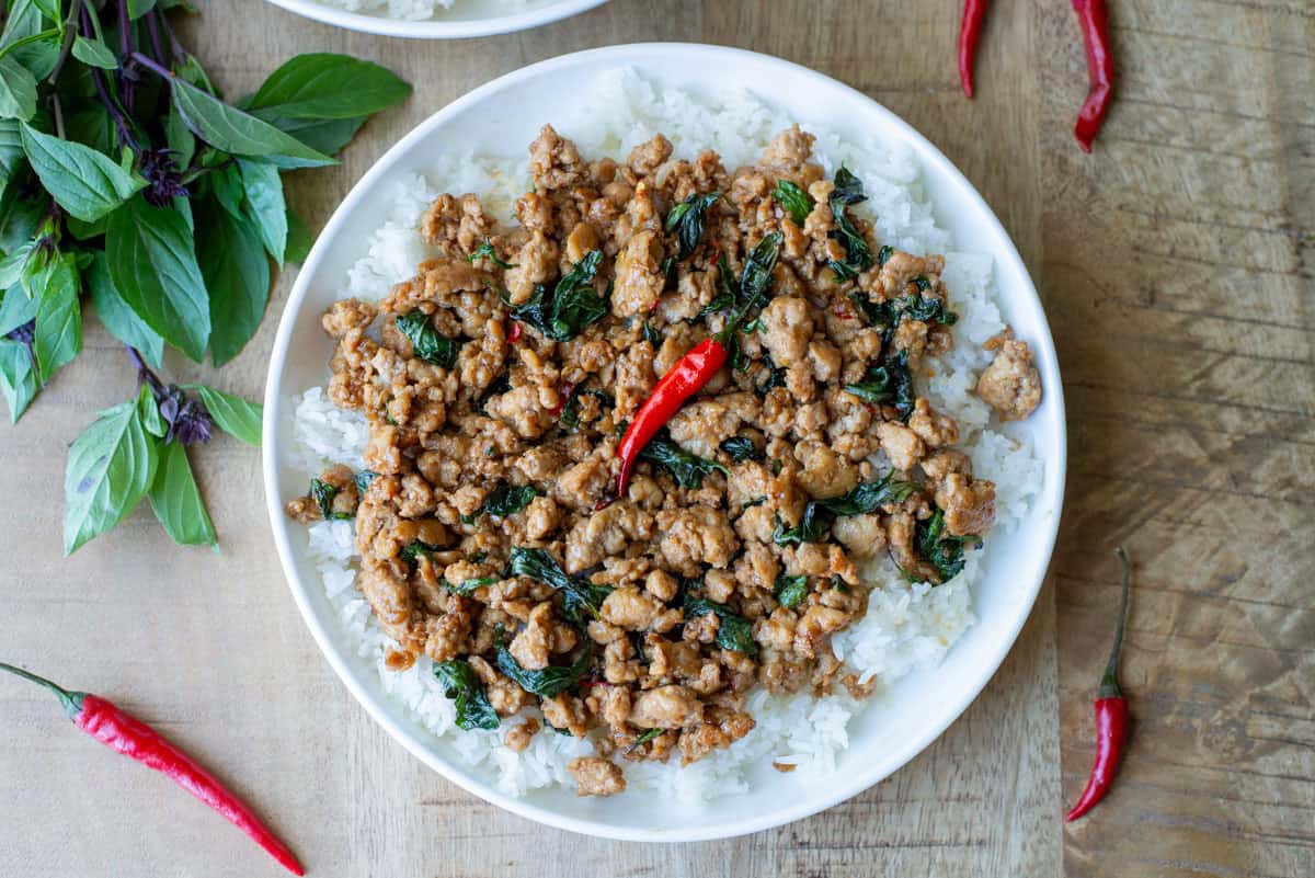 Overhead view of a full plate of Thai Basil Chicken over a bed of white rice with Thai Basil and Thai red chilies scattered on the table