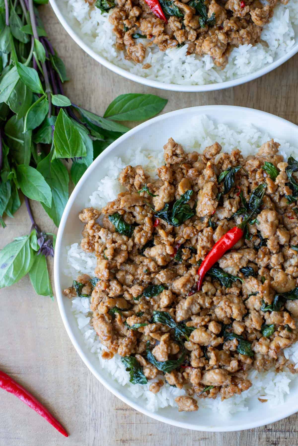 Overhead view of a partial plate of Thai Basil Chicken over a bed of white rice with another plate of Thai Basil chicken and Thai basil and Thai red chilies scattered on the table