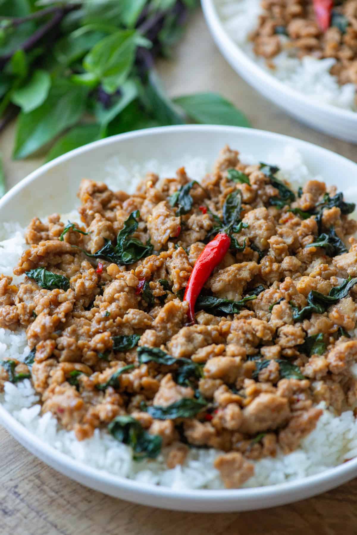 Overhead view of a partial plate of Thai Basil Chicken over a bed of white rice with another plate of Thai Basil chicken and Thai basil scattered in the background