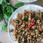 Overhead view of a partial plate of Thai Basil Chicken over a bed of white rice with another plate of Thai Basil chicken and Thai basil and Thai red chilies scattered on the table with a title overlay that says Thai Basil Chicken