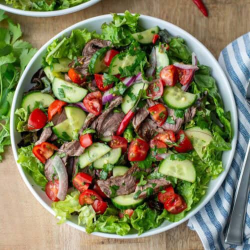Overhead view of plate of Thai beef salad over a bed of red leaf lettuce with another partial plate, cilantro stems, a napkin and a fork surrounding the plate