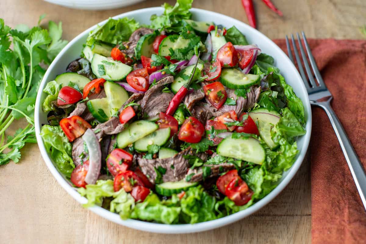 Overhead angled view of plate of Thai beef salad over a bed of red leaf lettuce with cilantro leaves, a napkin and a fork on the sides of the plates