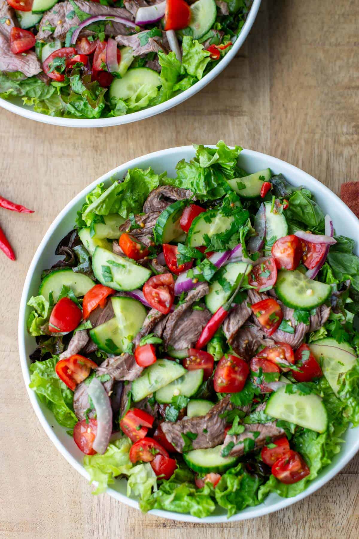 Overhead view of plate of Thai beef salad over a bed of red leaf lettuce with another partial plate in the background with a title overlay that says Thai Beef Salad