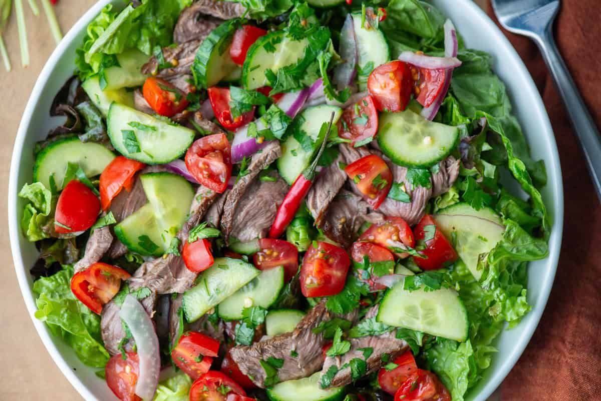 Overhead view of plate of Thai beef salad over a bed of red leaf lettuce with cilantro stems, a napkin and a fork on the sides of the plates