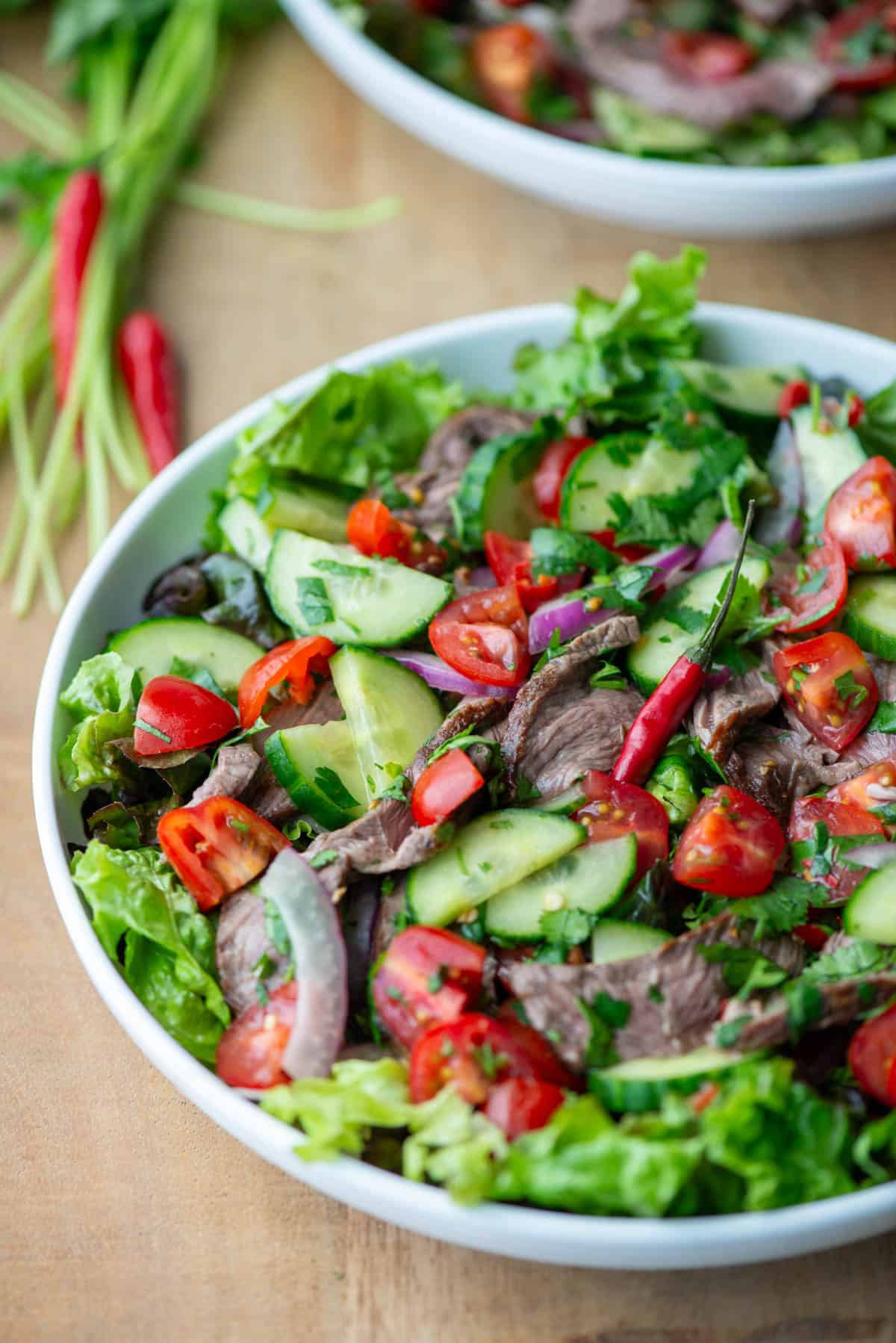 Overhead angled view of plate of Thai beef salad over a bed of red leaf lettuce with another partial plate, cilantro stems and Thai red chilies in the background