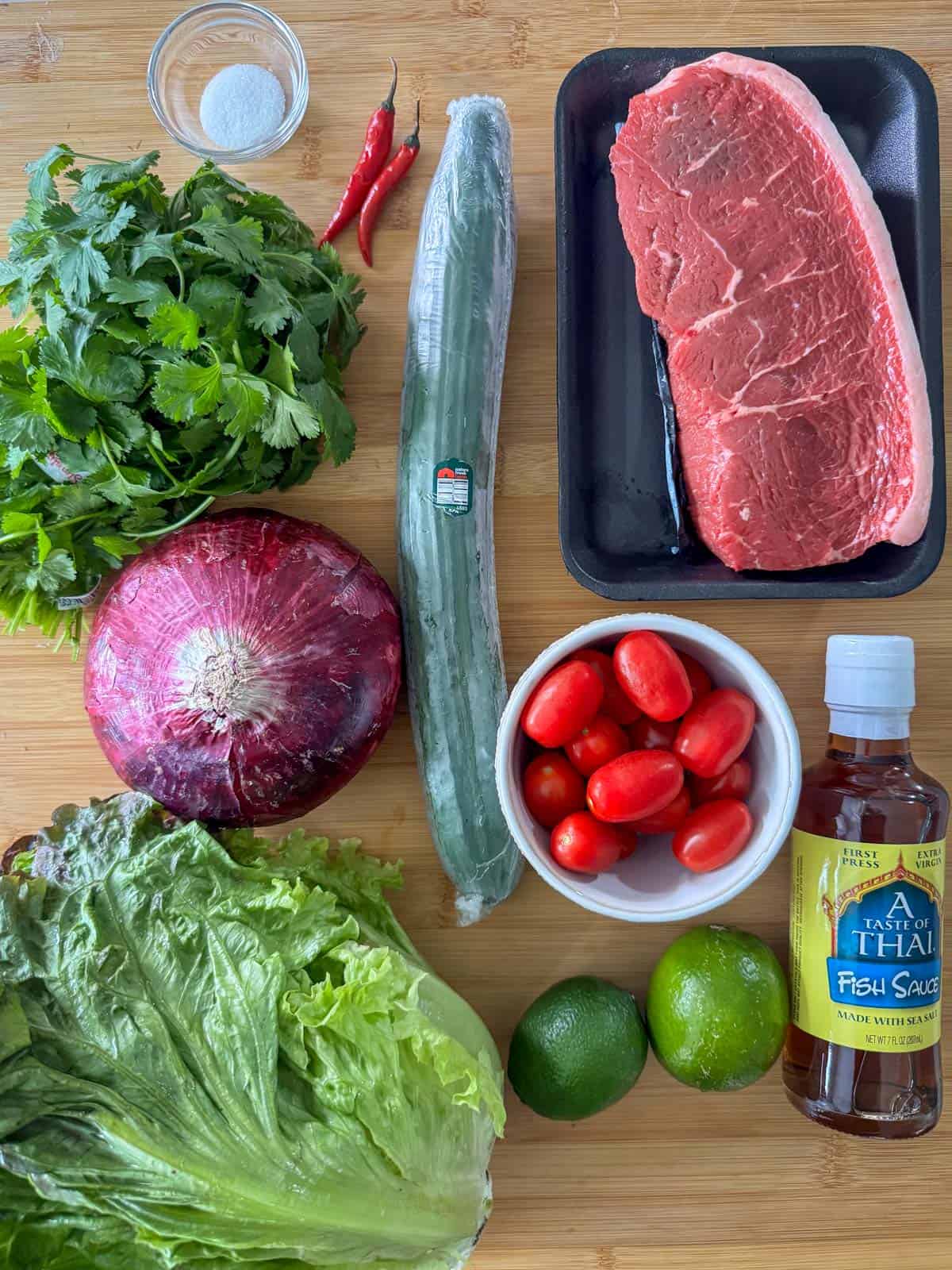 Ingredients needed for Thai beef salad: top sirloin steak, fish sauce, lime juice, Thai red chili peppers, white sugar, English cucumber, cherry tomatoes, red onion and cilantro to serve over red leaf lettuce.