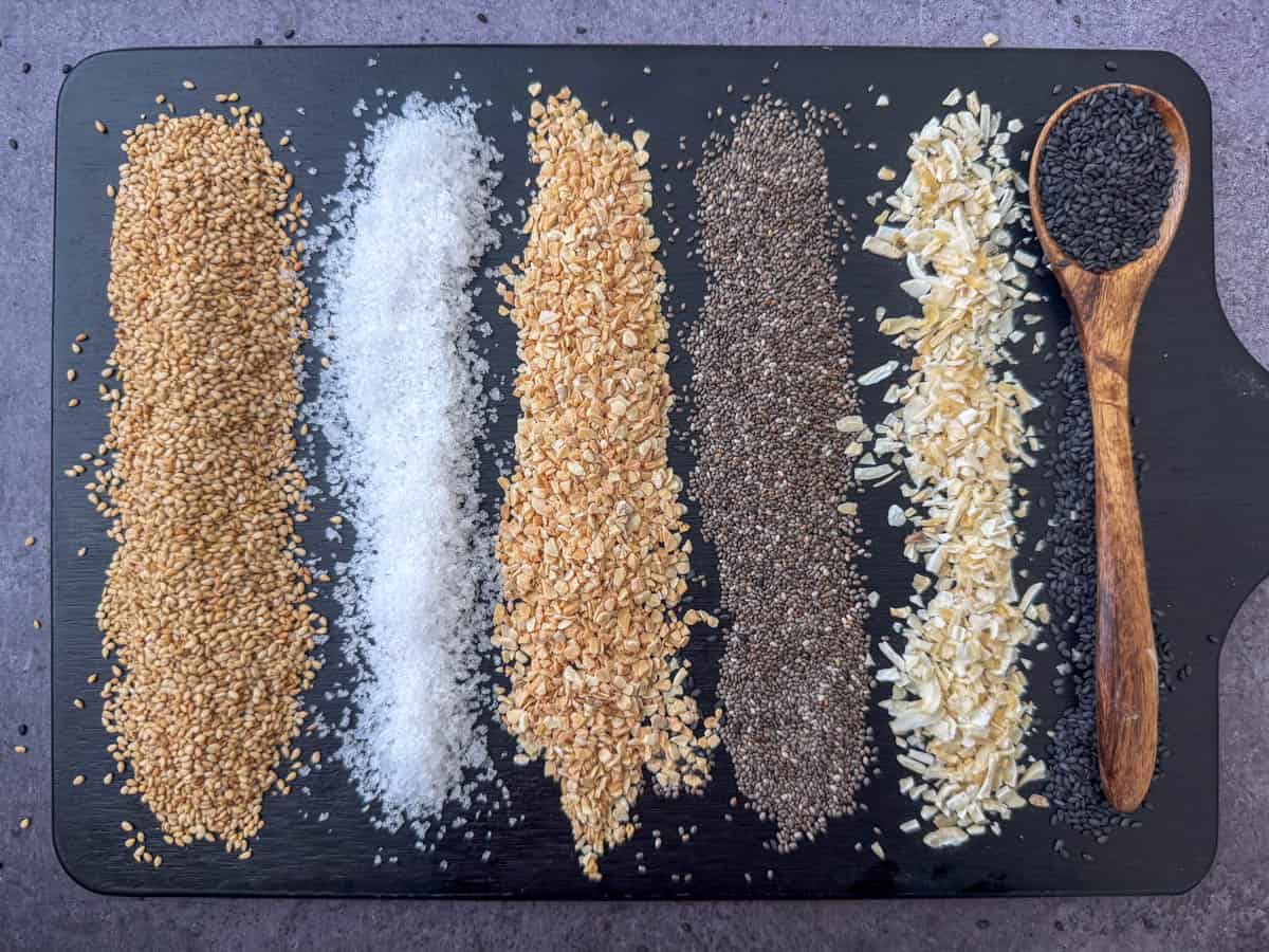 Spices needed to make everything but bagel seasoning each lined up on a cutting board: toasted white sesame seeds, flaked salt, minced garlic, poppy seeds, minced onion and black sesame seeds