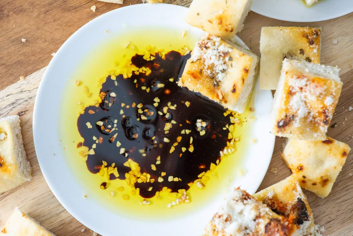 Overhead view of bread dipping oil on a small white plate with a cube of bread in the oil and more cubes of bread to the side of the plate