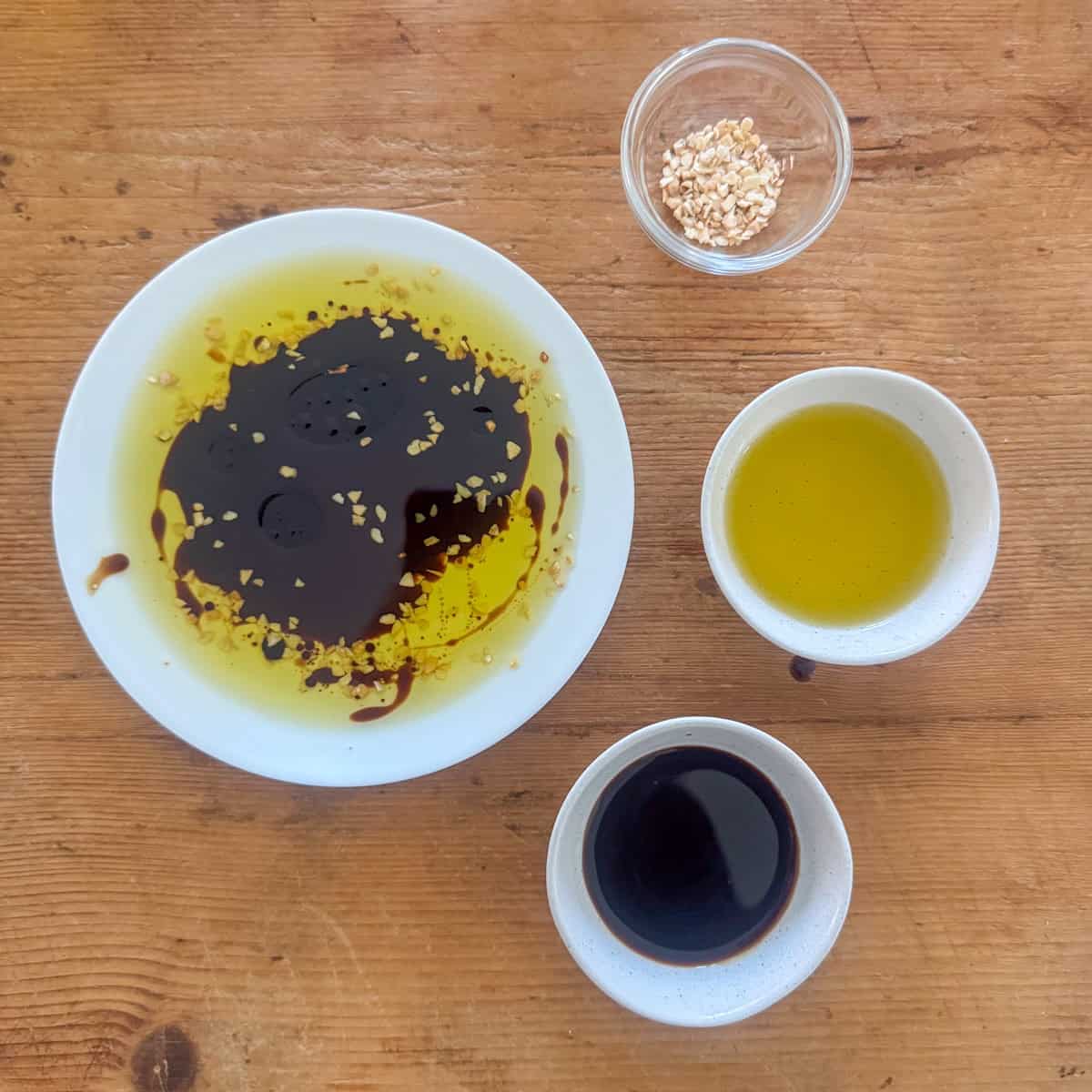 Overhead view of a plate of dipping oil next to three small bowls that contain the ingredients needed to make it: dried minced garlic, olive oil and balsamic vinegar