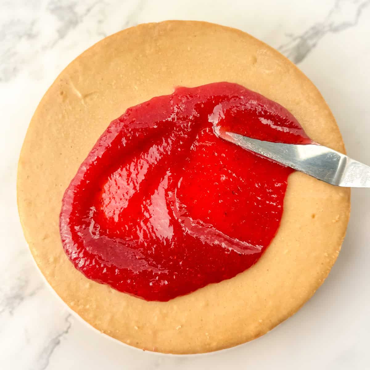 Overhead view peanut butter and jelly cheesecake with the jelly being spread on top of the peanut butter cheesecake