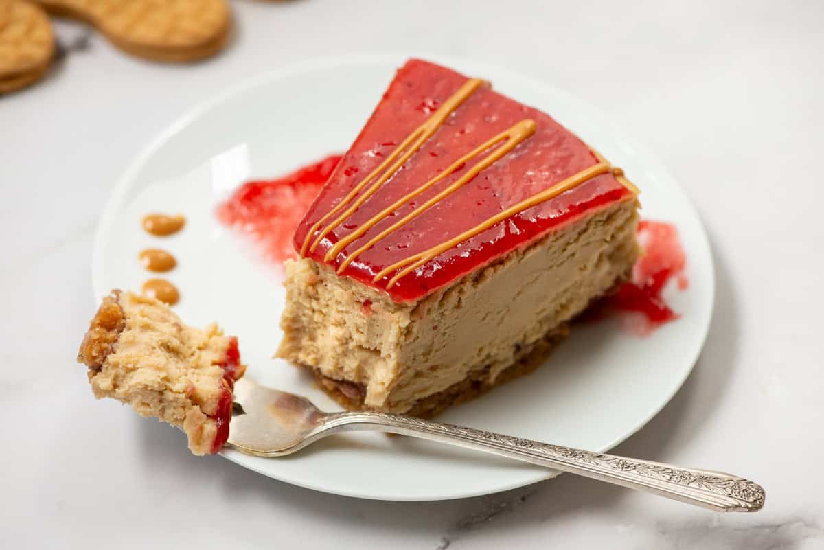 Side view of a slice of peanut butter and jelly cheesecake with peanut butter drizzle on a plate with a fork and a bite taken out