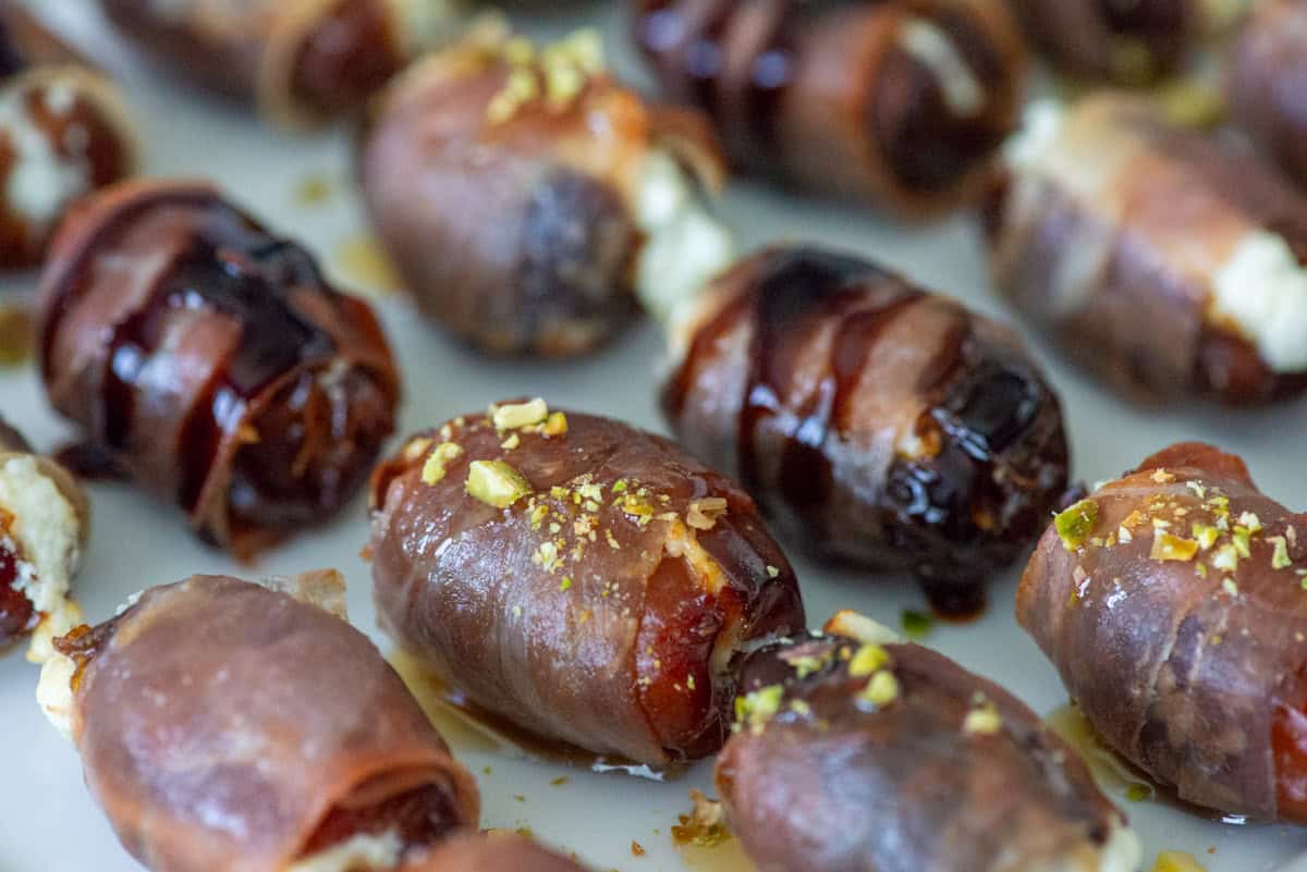 Overhead view of prosciutto wrapped dates at an angle on a white plate, some topped with balsamic glaze and some topped with hot honey and chopped pistachios